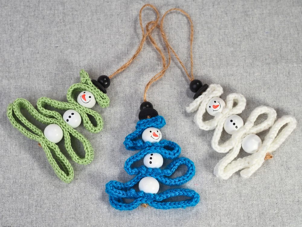 Three crochet snowman Christmas tree ornaments in white, blue, and green on a grey linen background.