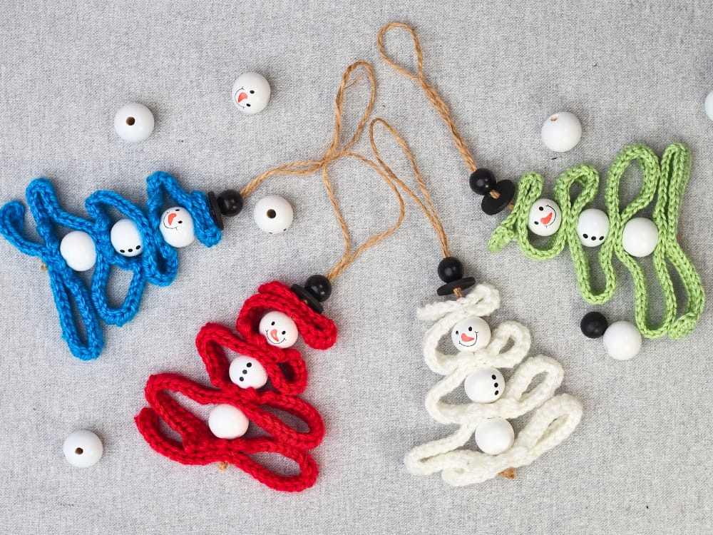 Four crochet snowman Christmas tree ornaments in red, white, blue, and green on a grey linen background.