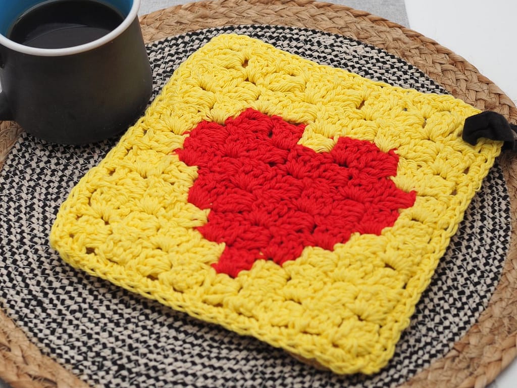 Finished C2C heart crochet potholder displayed on a woven placemat beside a coffee mug.