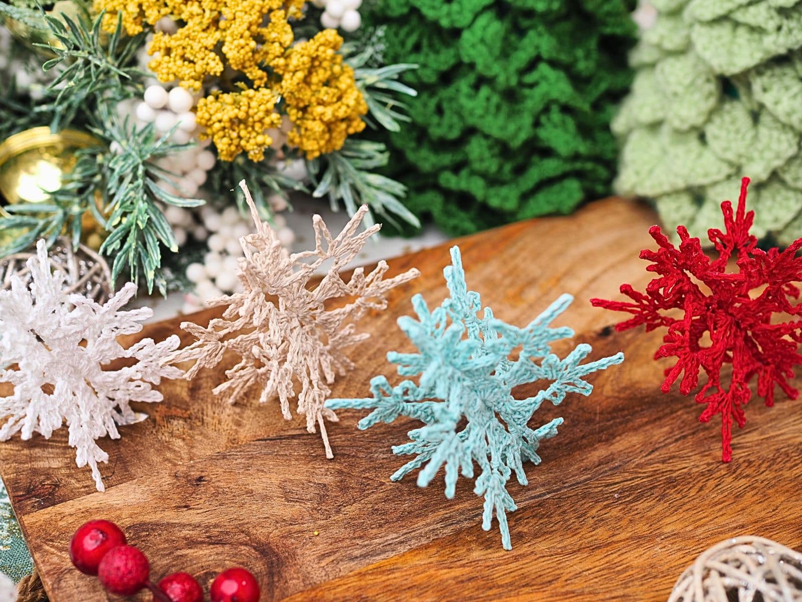 Four colorful 3D crochet snowflakes standing on a wood surface with Christmas decorations in the background.